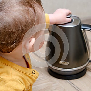 Toddler baby boy holds a hot kettle with boiling water. Child safety issues in the home room, little kid