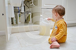 Toddler baby boy is playing with detergents and cleaning products in an open kitchen cabinet. Child safety issues in the home room