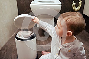 Toddler baby boy is playing in the toilet room with a trash can. Child plays on a brown floor in a beige bathroom with a trash can