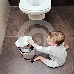 Toddler baby boy is playing in the toilet room with a trash can. Child plays on a brown floor in a beige bathroom with a trash can