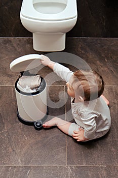 Toddler baby boy is playing in the toilet room with a trash can. Child plays on a brown floor in a beige bathroom with a trash can