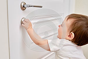 Toddler baby opens the lock, child hand close-up. White wooden door,