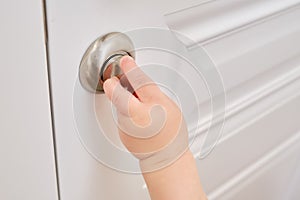 Toddler baby opens the lock, child hand close-up. White wooden door,