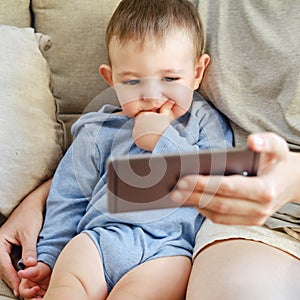 Toddler baby sits with his mother and looks into the phone. Mom and c