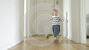 Toddler boy as he plays with a ball on the wooden floor of a long corridor in his house