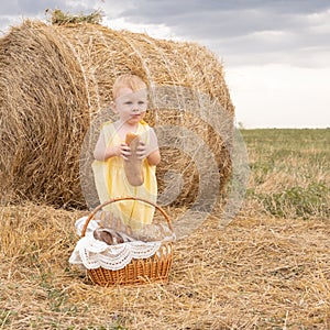 Toddler boy blond with basket of bread stands in a field with a haystack eating a baguette.