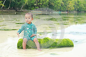 Toddler boy in blue swimsuit sitting on green trunk in shallow sea