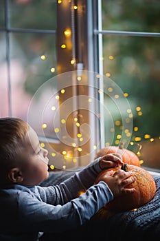 Toddler boy holding orange pumpkin on gray knitted plaid near window in evening surrounded with warm garland lights with golden