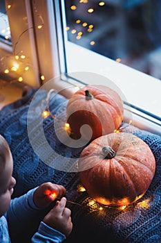 Toddler boy holding orange pumpkin on gray knitted plaid near window in evening surrounded with warm garland lights with golden