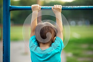 Toddler boy learning to do pull-ups exercise: preschooler hanging on worn-out steel bar outdoors, view from behind