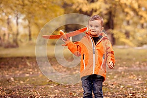 Toddler boy playing solo with orange toy airplane in autumn park. Trees with yellow and brown leaves in background