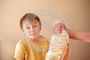 Toddler boy with puzzled face standing near sleeping newborn baby. Small boy meeting infant brother or sister and feeling confused