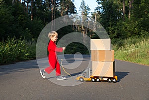 Toddler boy in red jumpsuit runs towards a big toy car - a truck with large cardboard boxes - parcels