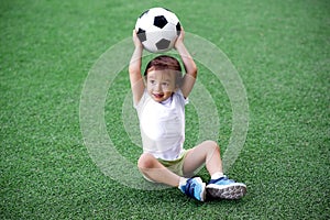 Toddler boy in sports uniform sitting on green footall field holding soccer ball above head. Copy space