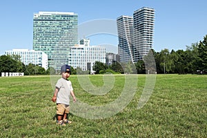 Toddler boy standing on green meadow in front of office buildings