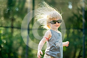 Toddler boy with sunglasses and messy hair jumping on a trampoline in a backyard. Sports and exercises for children. Summer