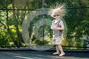 Toddler boy with sunglasses and messy hair jumping on a trampoline in a backyard. Sports and exercises for children. Summer