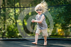 Toddler boy with sunglasses and messy hair jumping on a trampoline in a backyard. Sports and exercises for children. Summer