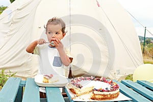 Toddler boy eating cheesecake with fruits and chocolate in front of tent