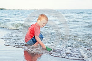 toddler child boy putting green paper boat in water on lake sea ocean shore 