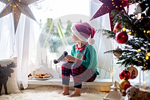 Toddler child, cute blond boy, sitting on the window in pajama, looking out for Satna Claus, Christmas lights around him
