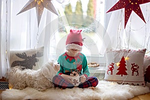 Toddler child, cute blond boy, sitting on the window in pajama, looking out for Satna Claus, Christmas lights around him