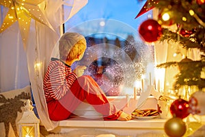 Toddler child, cute blond boy, sitting on the window in pajama, looking out for Satna Claus, Christmas lights around him