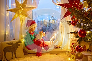 Toddler child, cute blond boy, sitting on the window in pajama, looking out for Satna Claus, Christmas lights around him