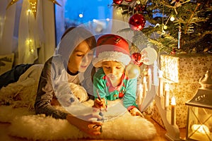 Toddler child, cute blond boy, sitting on the window in pajama, looking out for Satna Claus, Christmas lights around him