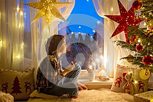 Toddler child, cute blond boy, sitting on the window in pajama, looking out for Satna Claus, Christmas lights around him