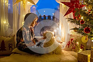 Toddler child, cute blond boy, sitting on the window in pajama, looking out for Satna Claus, Christmas lights around him