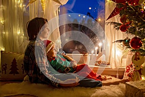 Toddler child, cute blond boy, sitting on the window in pajama, looking out for Satna Claus, Christmas lights around him