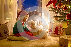 Toddler child, cute blond boy, sitting on the window in pajama, looking out for Satna Claus, Christmas lights around him