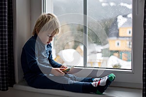 Toddler child, sitting on the window, watching the snow falling, reading a book