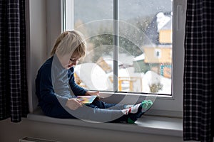 Toddler child, sitting on the window, watching the snow falling, reading a book