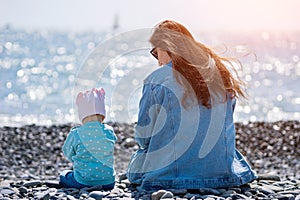 Toddler daughter and mother sit on sunlit pebble sea beach