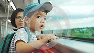 A toddler gazes out a train window, under the watchful eye of his mother