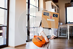 A toddler girl with carboard monster on head playing indoors at home.