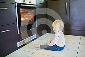 Toddler girl at home, sitting near the oven and waiting for food to be ready