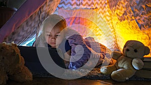 Toddler girl lies on blanket in playhouse watching cartoon on tablet with interest