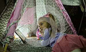 Toddler girl lies on blanket in playhouse watching cartoon on tablet with interest