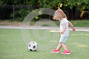 Toddler girl in pink sneakers playing football at stadium. Little blonde child preparing to kick soccer ball outdoors