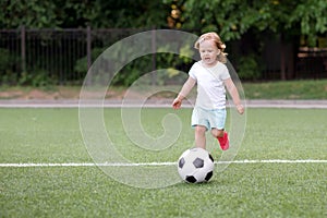 Toddler girl playing football: blonde child running on stadium to kick soccer ball. Outdoors activity and sports games concept