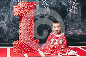 Toddler girl in a red dress in a white circle sitting near a big figure one. One-year-old girl on her birthday