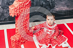 Toddler girl in a red dress in a white circle sitting near a big figure one. One-year-old girl on her birthday