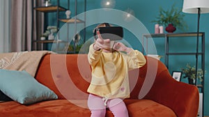 Toddler girl sitting on home sofa using virtual reality headset helmet app to play simulation game