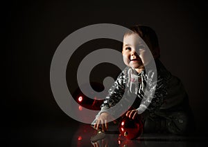 Toddler in gray boots and sparkling suit. She playing with three red balls, sitting on floor. Twilight, black background. Close up