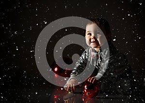Toddler in gray boots and sparkling suit. She playing with three red balls, sitting on floor. Twilight, black background. Close up