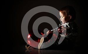 Toddler in gray boots and sparkling suit. She playing with three red balls, sitting on floor. Twilight, black background. Close up