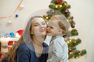 Toddler kisses mom on the cheek against the background of the Christmas tree and fireplace